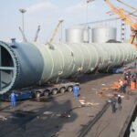 Large grey industrial process column transported horizontally on modular trailers beside dockside cranes.